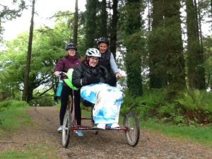 Student with two staff on adaptable bicycle in woods